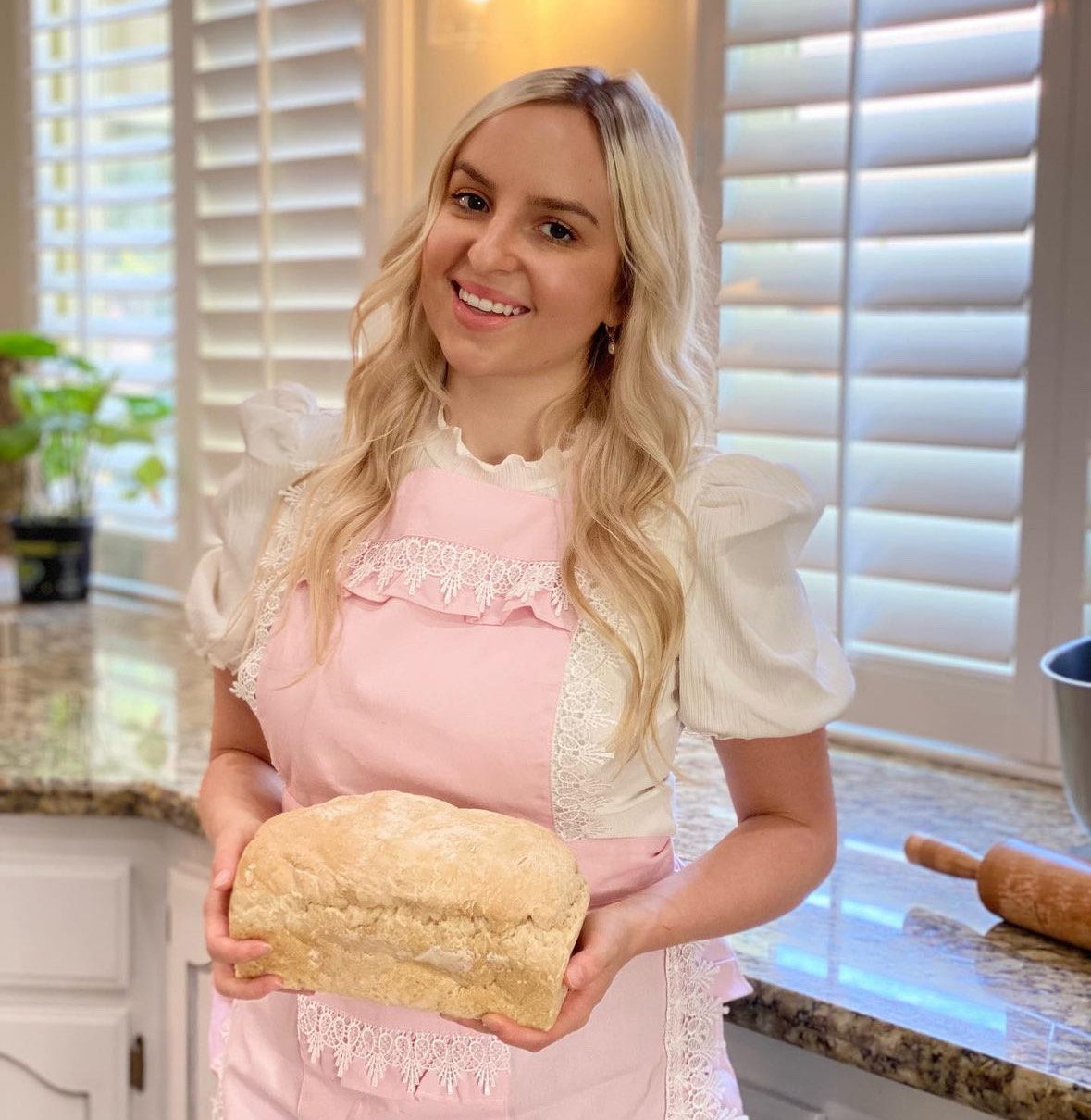 Young woman with long blonde hair wearing a pink apron, holding a loaf of freshly baked sourdough bread.